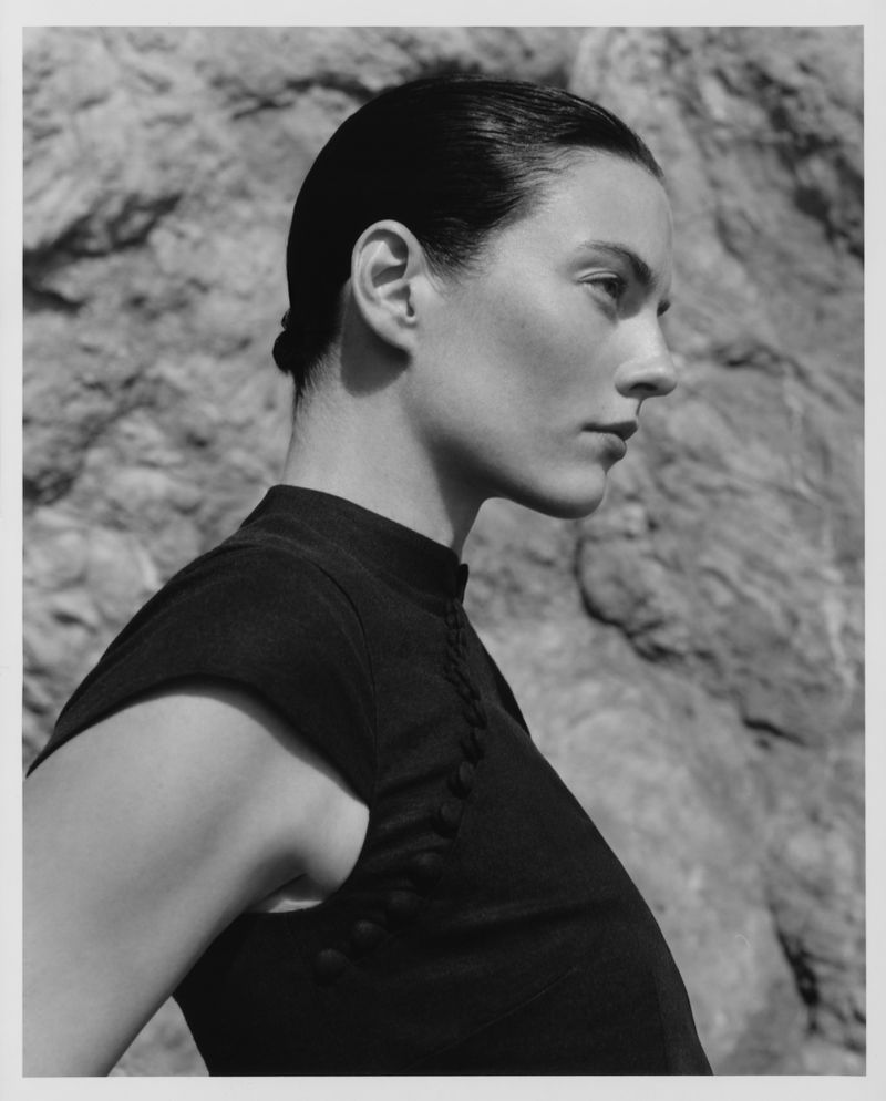 Profile portrait of a model with hair tied back wearing a black sleeveless top against a rocky textured background.