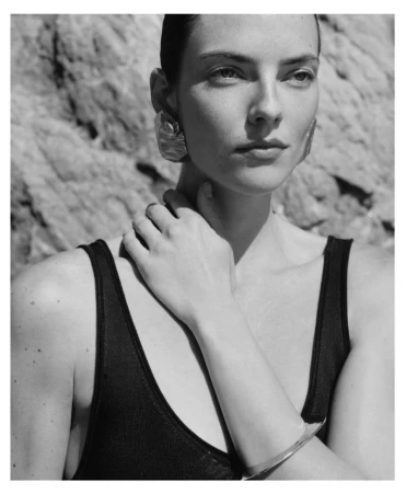 Model wearing a black tank top and statement earrings posing against a rocky coastal backdrop outdoors.