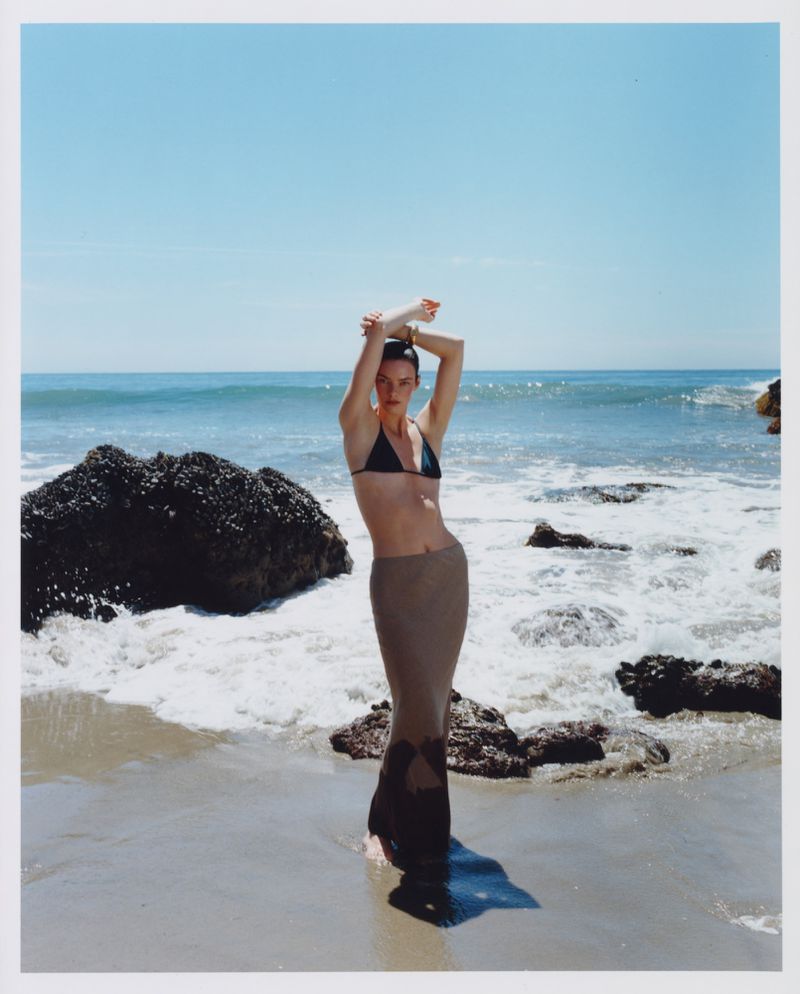 Model wearing a dark bikini top and long skirt posing on a sandy beach with ocean waves in the background.
