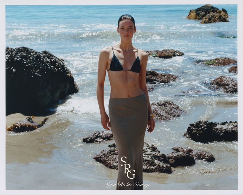 A woman in a bikini and skirt poses on a beach with rocks and waves. The image captures a natural, sunny beach scene with clear ocean water.