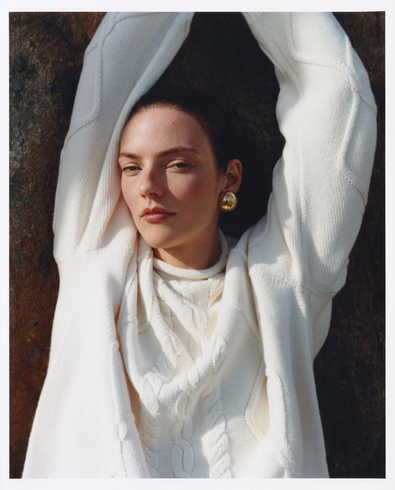 Model wearing a white cable knit sweater with arms raised against a textured dark background for Revolve.