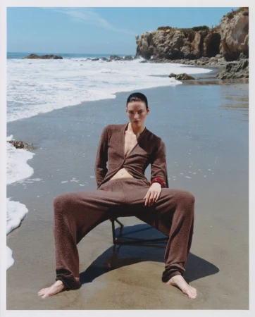 Model seated in a chair on a sandy beach wearing a brown knitted two-piece outfit with ocean waves and rocky cliffs behind.