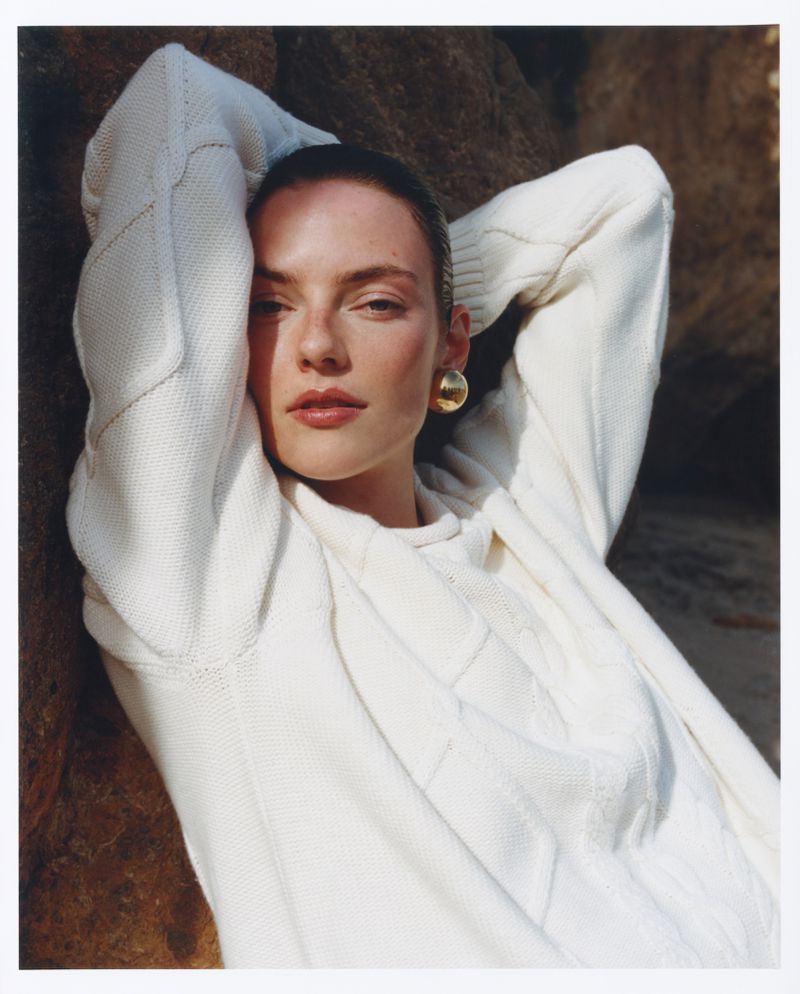 Model wearing a white cable-knit sweater with hands behind her head against a rocky outdoor background for Revolve.