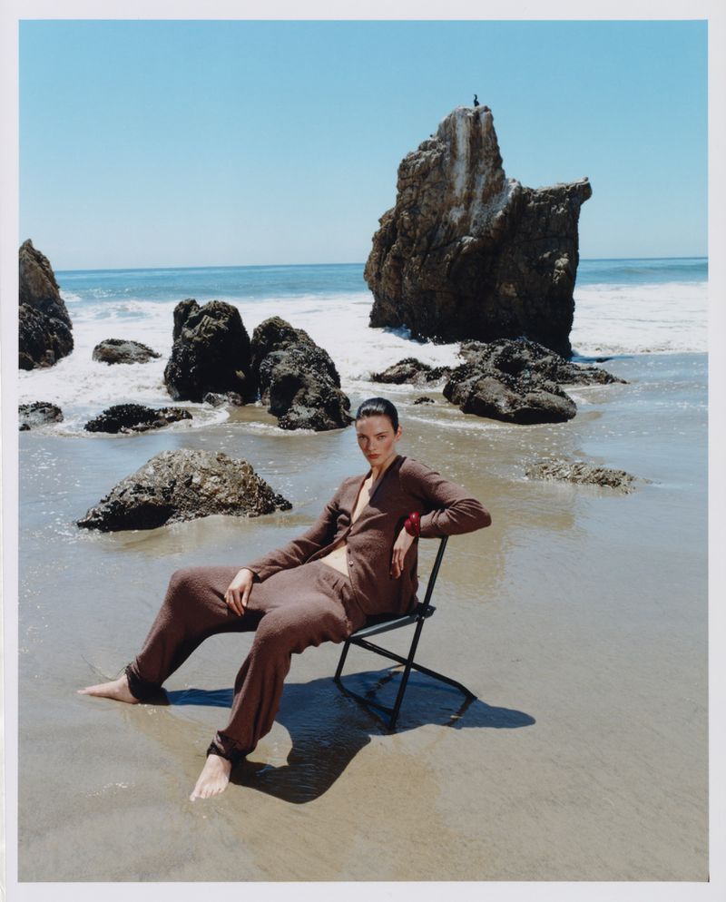 Model seated on a folding chair on a wet sandy beach wearing a brown knit co-ord set with rocky outcrops in the background.