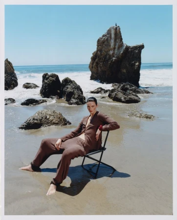 Model seated on a folding chair on a wet sandy beach wearing a brown knit co-ord set with rocky outcrops in the background.