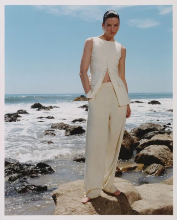 Model standing on rocky coastal shore wearing cream sleeveless top and matching trousers under blue sky
