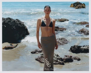 A female model standing on a rocky beach wearing a dark bikini top and long skirt for a Revolve fashion campaign.