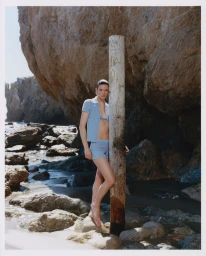 Model leaning against a weathered wooden post on a rocky beach wearing a matching blue polo and shorts set