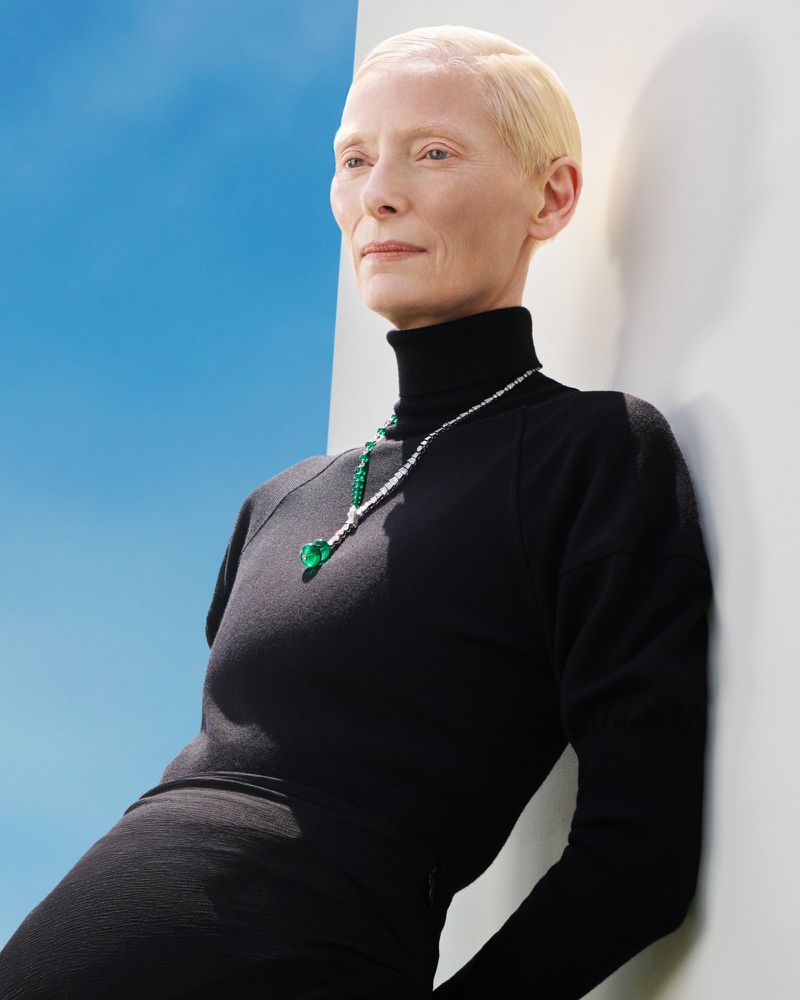 Tilda Swinton wearing a black turtleneck and fine emerald necklace leaning against a white wall under a blue sky.
