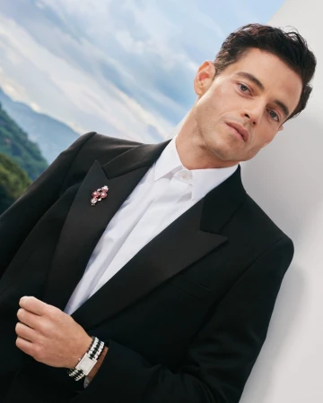 Portrait of Rami Malek in a black tuxedo, white shirt, and jeweled brooch, posing in front of a background featuring mountains and sky.