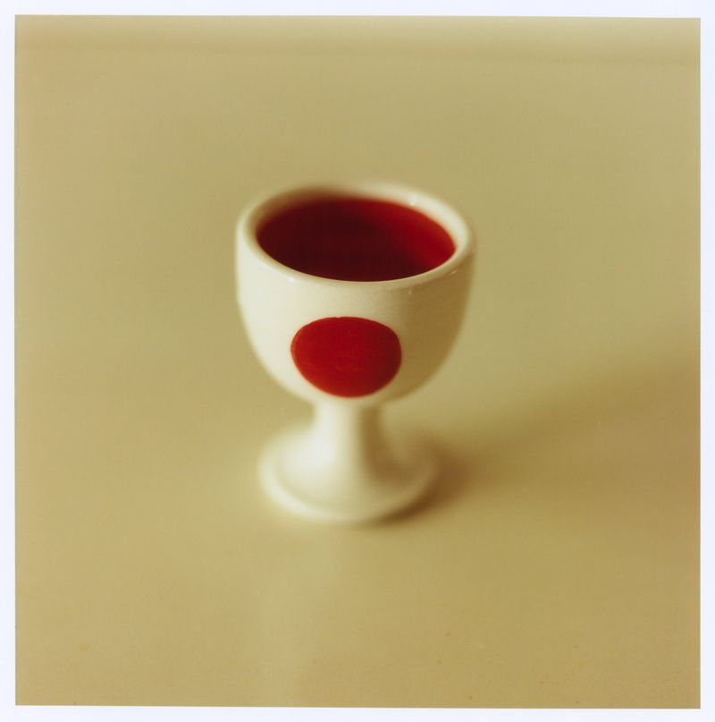 Close-up of a small white ceramic egg cup with a red polka dot on a cream surface, photographed on instant film.