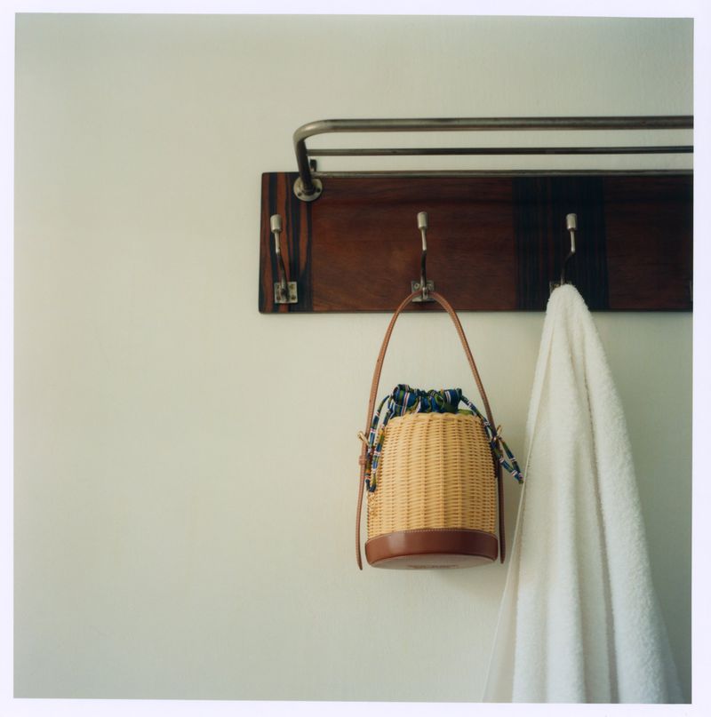 Woven straw bucket bag with leather base hanging from a wooden wall rack alongside a white towel.