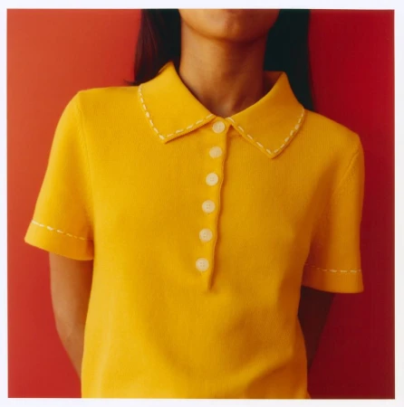 Close-up of a person wearing a bright yellow polo shirt with white stitch details against a solid red wall.