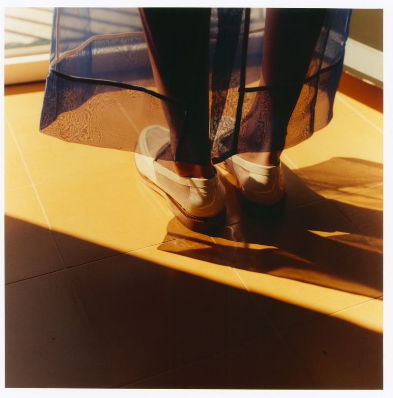 Close-up of legs wearing a translucent blue skirt and white loafers standing on a yellow tiled floor in sunlight.