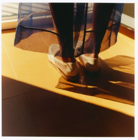 Close-up of legs wearing a translucent blue skirt and white loafers standing on a yellow tiled floor in sunlight.
