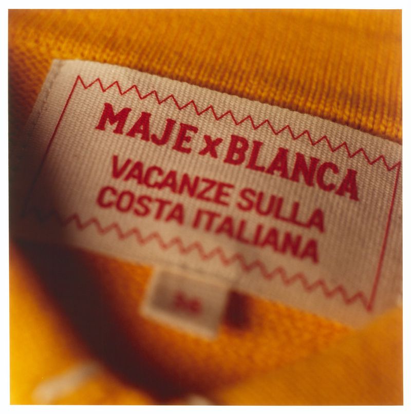 Close up of a clothing brand label reading MAJE x BLANCA and VACANZE SULLA COSTA ITALIANA sewn into yellow knitwear.