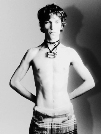 Monochrome portrait of a slender male model wearing a statement necklace and plaid pants in a neutral studio setting with soft light.