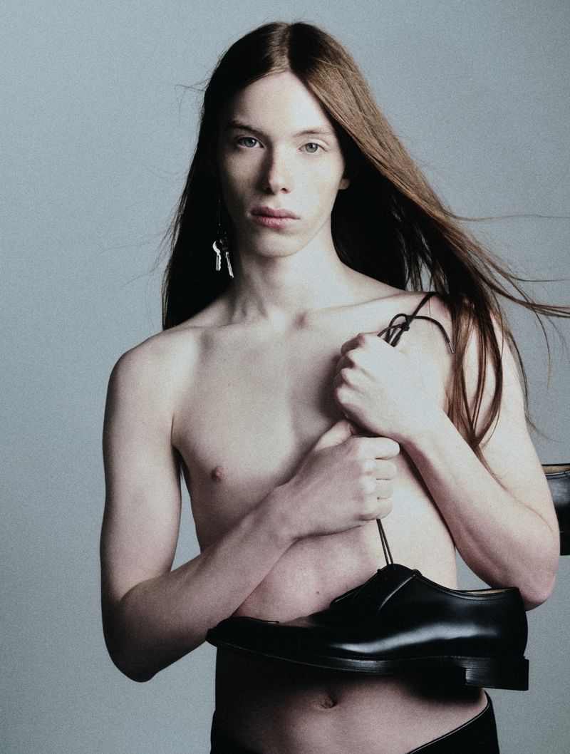 Nude male model with long hair poses with a black shoe in a studio setting. He is wearing a black bottom.