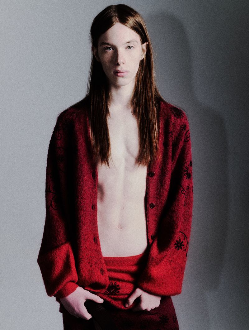 Portrait of a young adult man with long hair, wearing a patterned red cardigan, posing in a photo studio with neutral gray background.
