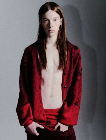 Portrait of a young adult man with long hair, wearing a patterned red cardigan, posing in a photo studio with neutral gray background.