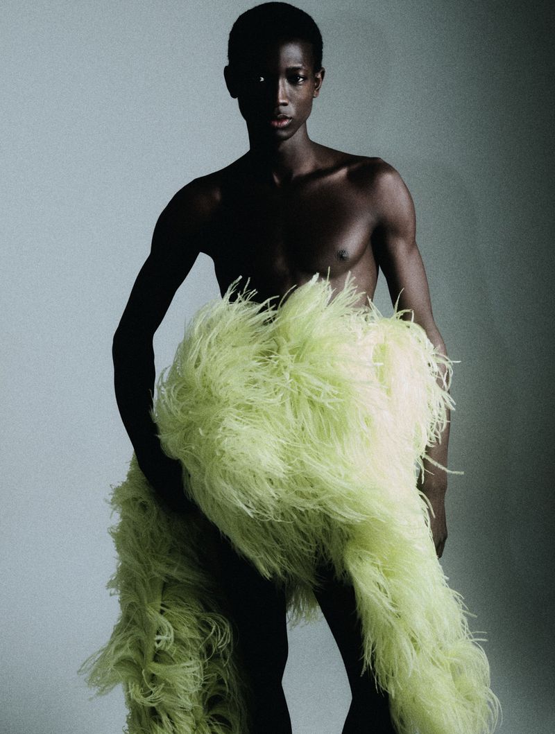 Striking portrait of a topless person with dark skin draped in vibrant green feathers, set against a soft grey background in a studio setting.