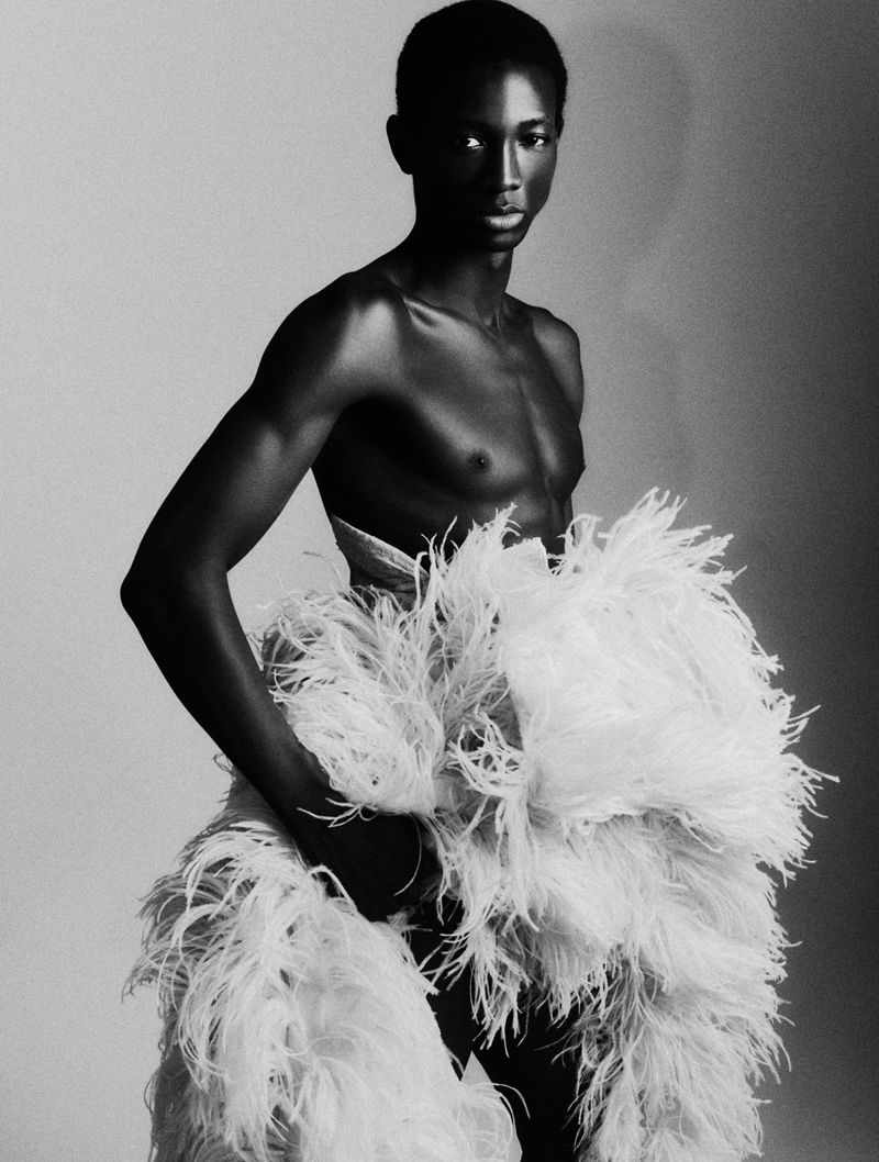 Striking black and white portrait of a shirtless African male model wearing a large white feathered skirt against a neutral backdrop.