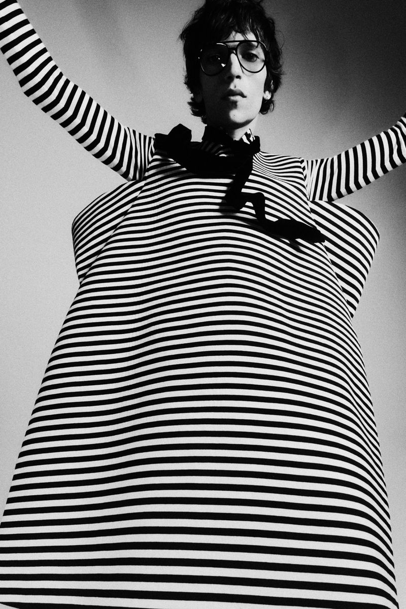 Black and white portrait of a person with glasses wearing a striped top and dress, arms raised, shot from a low angle.