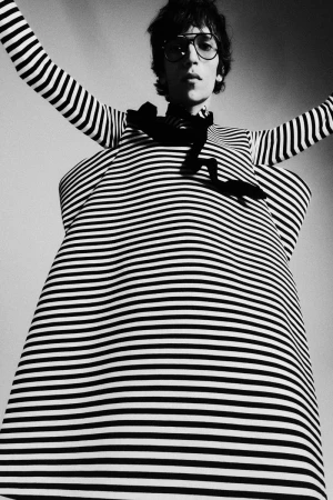 Black and white portrait of a person with glasses wearing a striped top and dress, arms raised, shot from a low angle.
