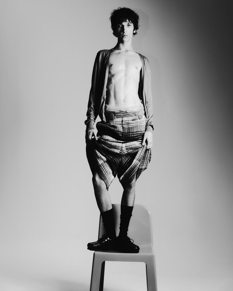 Young man with bare chest stands on a gray chair with black socks and loafers. He is wearing a cardigan and patterned skirt.