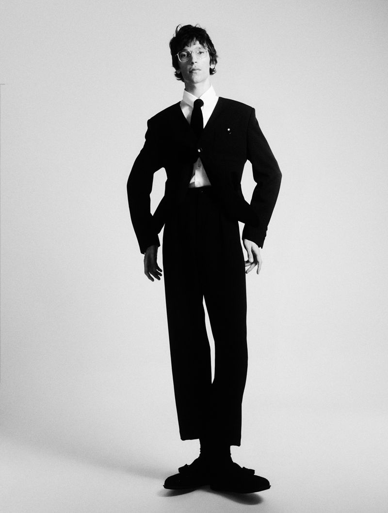 Elegant adult male in a black suit and tie poses in a full shot against a plain white backdrop, studio lighting, formal attire.