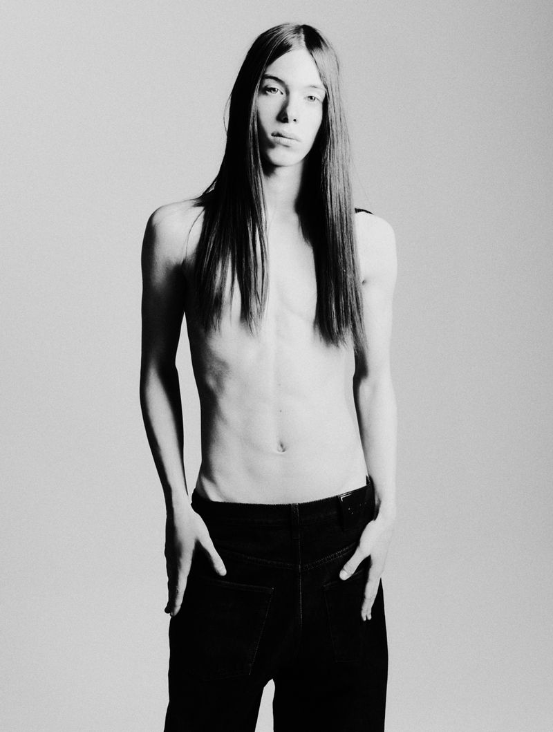 A slender, shirtless adult male stands in a studio with hands in his pockets, wearing black pants, and has long, straight brunette hair.