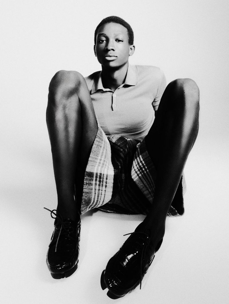 A young black adult poses with legs spread on the floor wearing shorts and a polo shirt in a studio with white background.