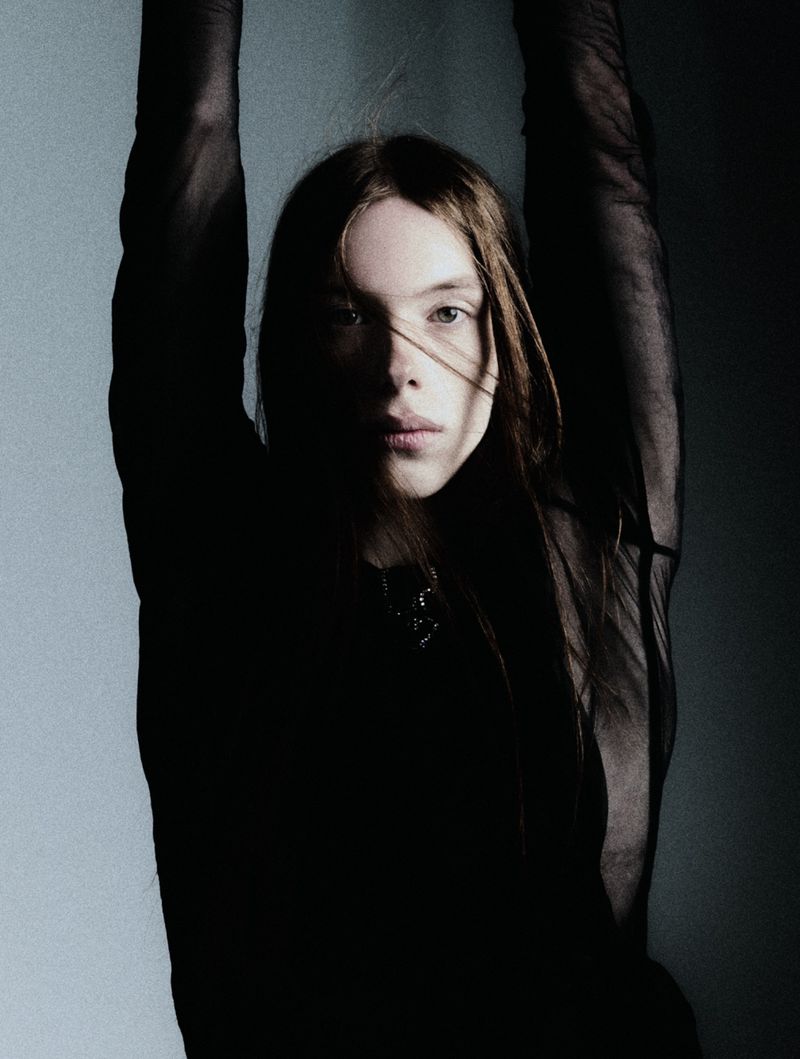 Portrait of a young woman with long brown hair in a black top, standing with arms raised against a gradient wall, in shadow and light.