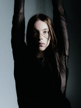 Portrait of a young woman with long brown hair in a black top, standing with arms raised against a gradient wall, in shadow and light.