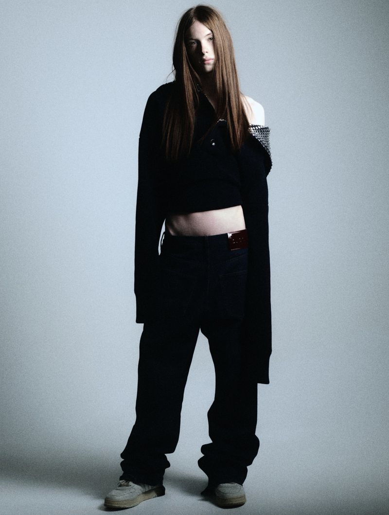 Full-length portrait of a woman with long brown hair, wearing a black top, loose black pants and casual shoes in a studio setting.