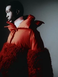 Black model wearing an orange puffer jacket with textured red faux fur cuffs against a grey studio background