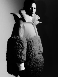 Black and white studio portrait of a Black male model wearing an oversized sculptural coat with voluminous textured shearling lower body.