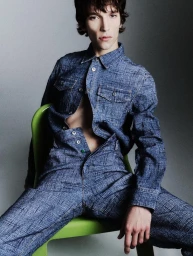 Model wearing a blue denim shirt and matching trousers seated on a bright green chair against a plain grey studio backdrop.