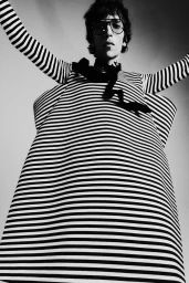 Low-angle shot of a male model in black and white striped voluminous clothing and glasses looking down against a studio backdrop.