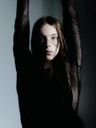 Model with arms raised wearing a black sheer top against a grey backdrop