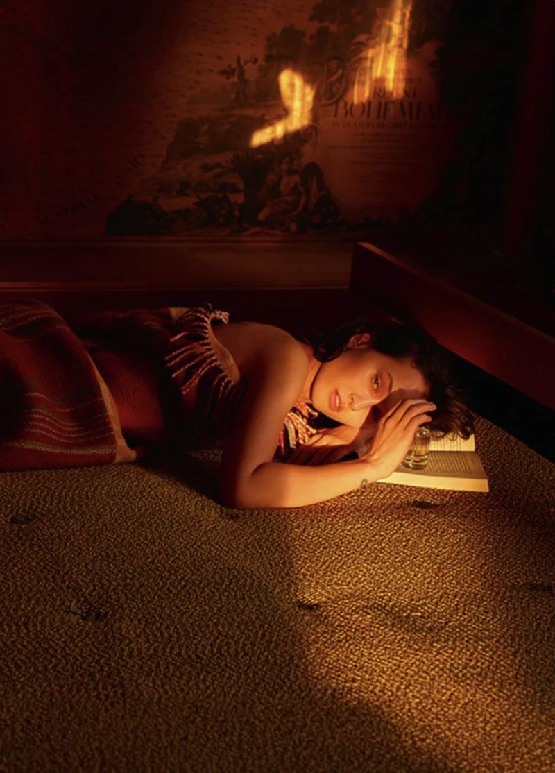 Woman lying down on a carpeted floor next to an open book and a small glass perfume bottle, illuminated in warm amber light.