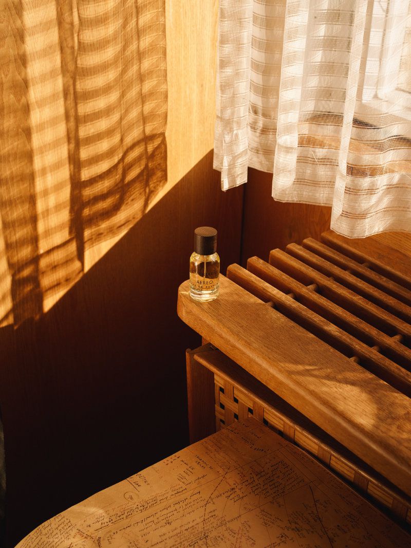 A perfume bottle standing on a wooden surface next to sheer white curtains with warm sunlight casting shadows