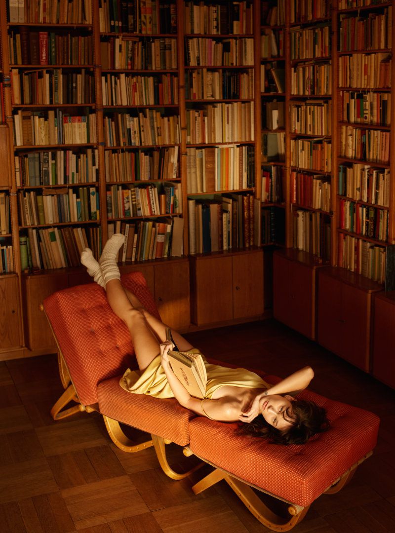 Woman in a gold dress reclines with a book in a library. Hardwood floors and floor-to-ceiling bookshelves filled with books.