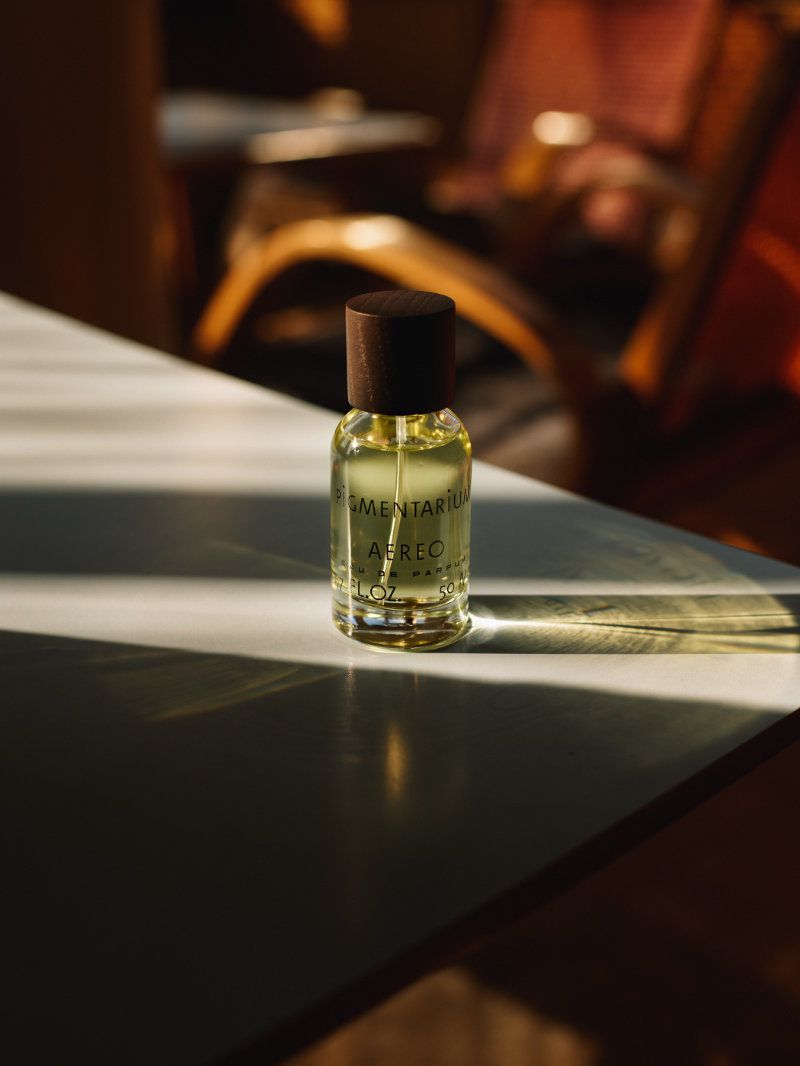 A small glass perfume bottle with a dark wooden cap standing on a table under warm sunlight, photographed by Hana Knizova.