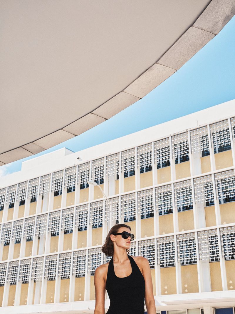 Model wearing dark sunglasses and a black halter top outdoors against a modern building with a concrete overhang