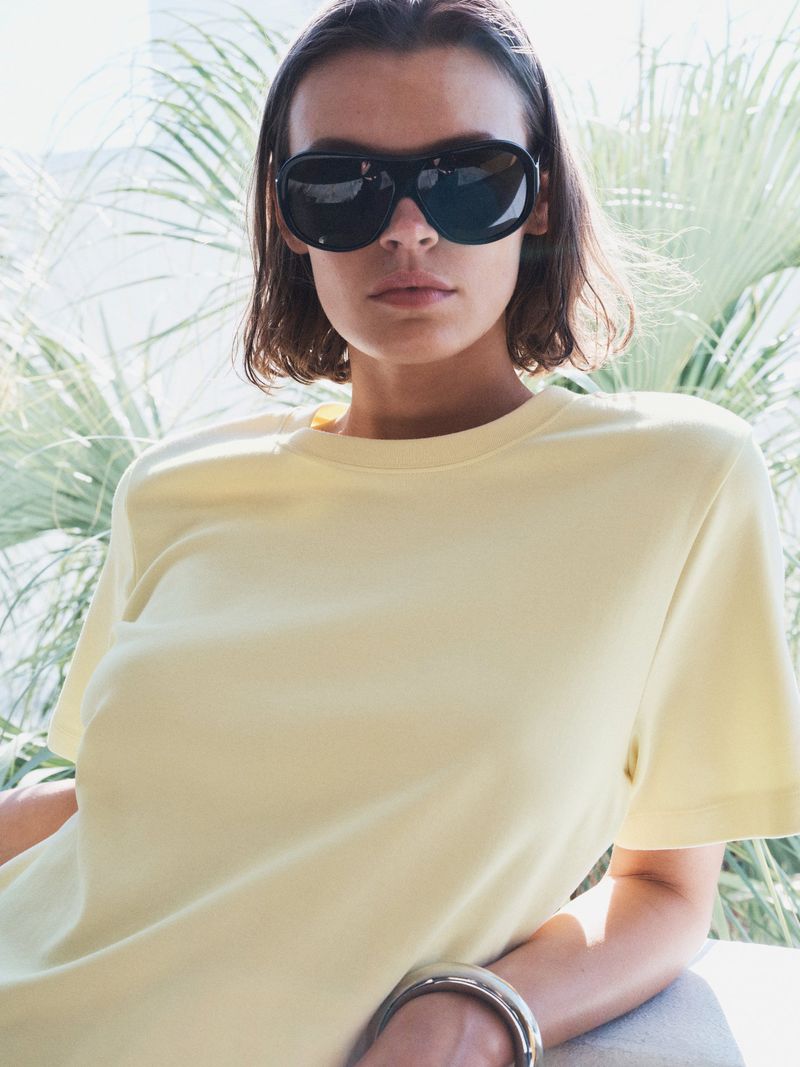 Cara Taylor wearing large black sunglasses and a yellow t-shirt outdoors with palm leaves in the background.