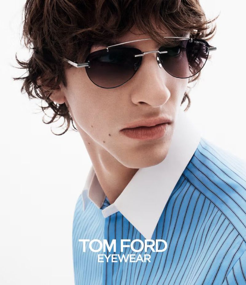 A light-skinned male model with wavy hair wears frameless sunglasses and a striped blue shirt with a white collar in a studio shot.