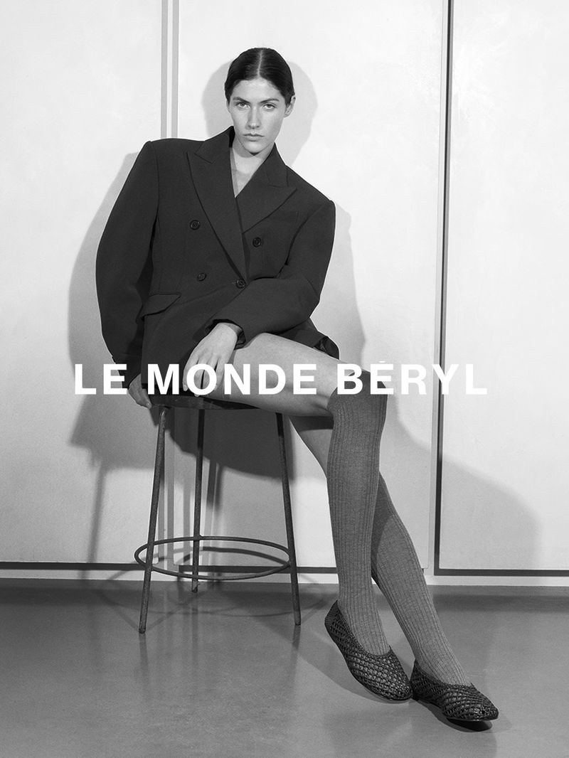 Black and white fashion shot of a woman wearing a blazer, knee-high socks, and woven shoes while sitting on a stool.