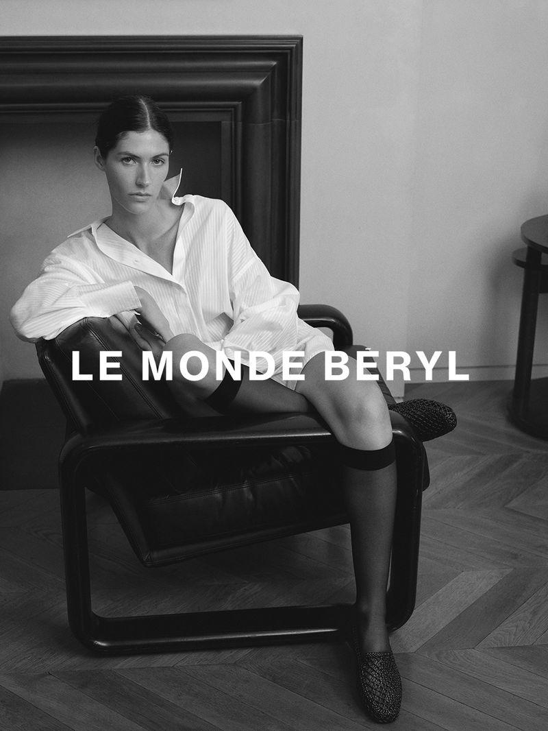 Monochrome fashion campaign image of a model seated in a leather armchair wearing an oversized white shirt and black flats.