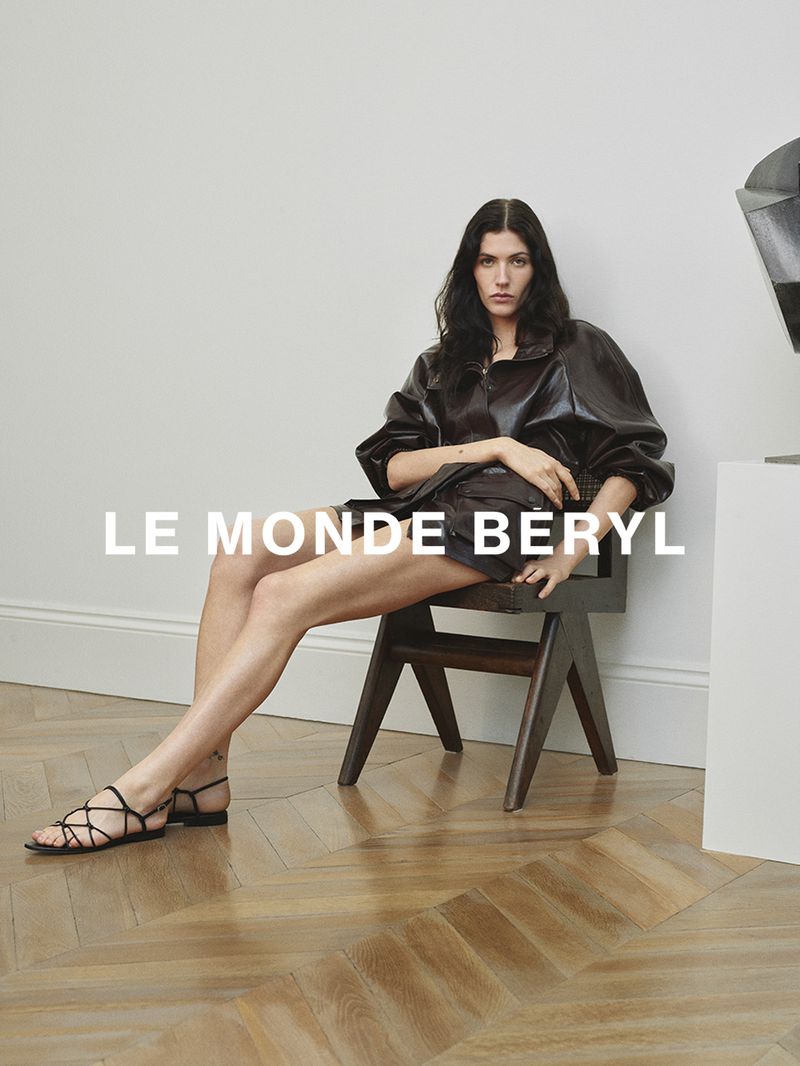 Model seated on a wooden chair wearing dark leather apparel and strappy flat sandals against a light wall